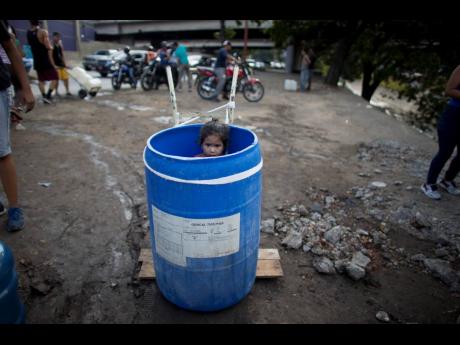 In this March 11, 2019, file photo, a little girl stands inside a plastic barrel while her family waits to collect water from an open pipe above the Guaire River, during rolling blackouts in Caracas, Venezuela.