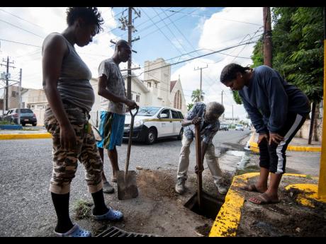 From left: Little North Street residents Sharon Tucker, Devon Myers and Verna Jones look on as 71-year-old Andral Johnson loosens the dirt and silt for removal during a drain-cleaning exercise at the intersection of North Street and Johns Lane in Kingston on November 26.