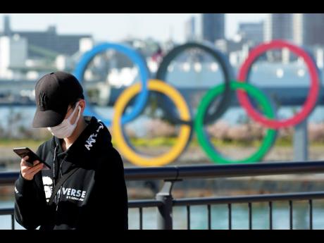 In this March 3, 2020 photo, a tourist wearing a protective mask takes a photo with the Olympic rings in the background, at Tokyo’s Odaiba district in Tokyo. 
