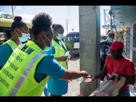 Public health aides sanitise the hands of a snacks vendor as they prepare to give her a mask outside the Coronation Market in downtown Kingston yesterday during a COVID-19 sensitisation and testing initiative.