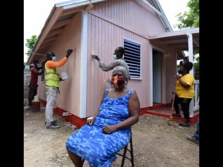 Joan Willie sits waiting in her Braeton Meadows yard in Portmore, St Catherine, yesterday as Food For The Poor workmen add the finishing touches to her house donated in partnership with Boom Energy Drink. Willie was selected for the house as the roof of he