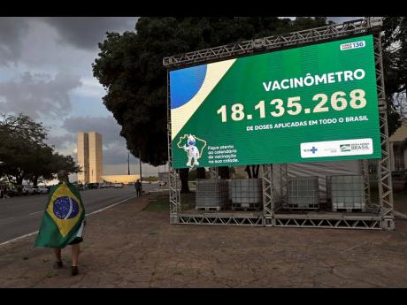 A Brazilian wearing the national flag walks by a billboard presenting the number of COVID-19 vaccines administered, in Brasilia, Brazil, on March 30.