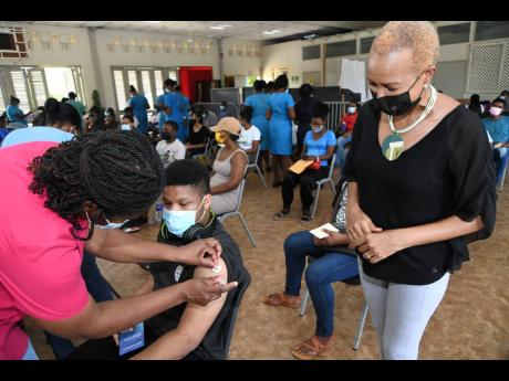 Education Minister Fayval Williams observes 16-year-old Matthew Reid of Wolmer’s Boys’ School getting his first dose of the Pfizer vaccine from midwife Naomi Baugh Brown during an inoculation blitz at Portmore HEART Academy on Sunday.