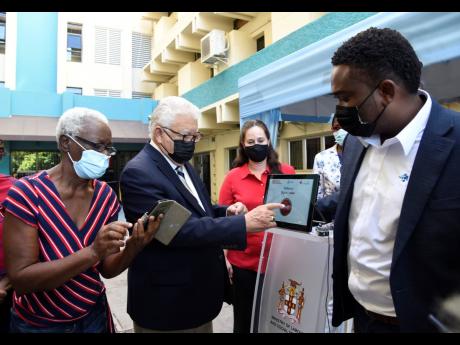 Lilieth Curtis (left), one of the beneficiaries of the COVID-19 Cash Assistance Programme, joins Labour and Social Security Minister Karl Samuda (second left) in tapping away to access cash under the welfare programme at the ministry’s North Street compo
