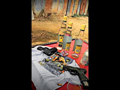 Two handguns, a Glock and Taurus, are displayed alongside rum and ammunition on a table with a memorial T-shirt bearing the image of slain reputed gunman Kirk ‘Big Red’ Wint at a wake in the Banga Gully area of Christian Pen last Saturday. Wint was sho
