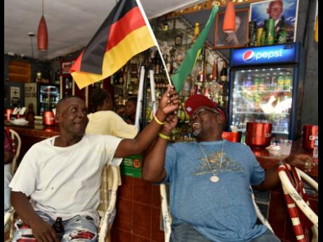 Germany supporter Martin Powell (left) and  Brazil fan ‘Blinds’ wave their teams’ flags while watching the United States-Wales match at the Movie Star lounge on Omara Road in Kingston.