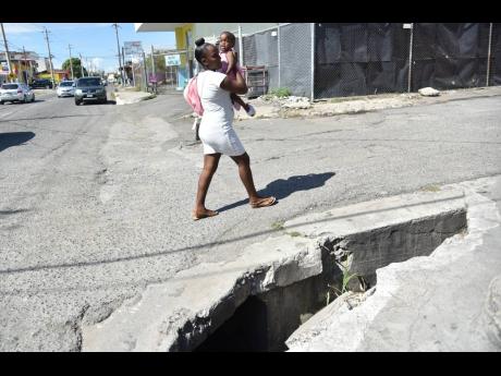 A mother and holds her child safely in her arms as they pass by an uncovered storm drain at the intersection of Homestead and Windward roads in Kingston on Tuesday. A visually impaired man became the latest victim to fall into the drain earlier this week, 