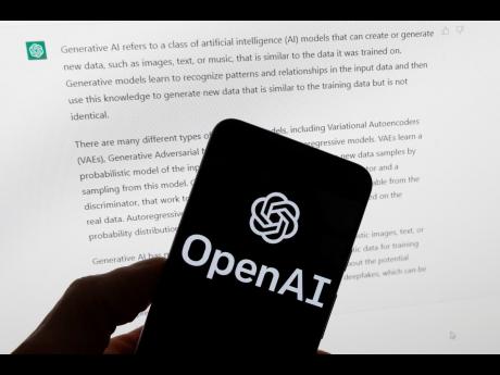 The OpenAI logo is seen on a mobile phone in front of a computer screen displaying output from ChatGPT.
