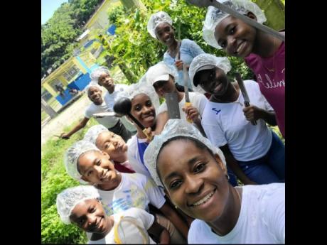 Aneta Blackett-Ricketts surrounded by young volunteers displaying kitchen utensils. The girls assist in cooking the meals and distributing them to needing persons. 