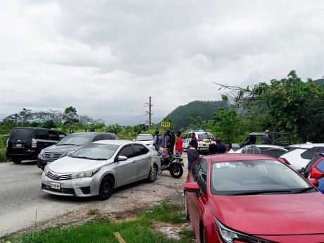 Members of the security forces and onlookers converge along the Eglinton main road at Spur Tree , Manchester, where the body and motor vehicle belonging to Reverend Orville Moore were found yesterday. Members of the security forces had launched a two-day s