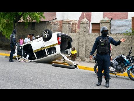 A policeman directs traffic past an overturned Toyota Probox taxi on North Street in KIngston yesterday. The driver of the  taxi was reported to have broken the stop light at the intersection of North and Duke streets and collided with a Honda Stream motor